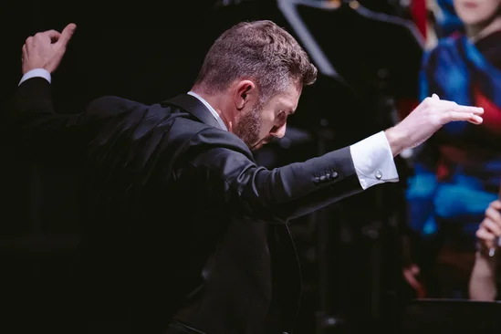 Conductor in a suit leading an orchestra, arms raised, focused expression.